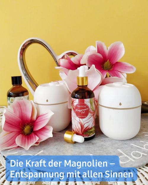 Nahaufnahme eines Stilllebens, das zwei weiße kerami­sche Diffusoren, zwei Flaschen ätherischer Öle (eine bernsteinfarbene und eine klare) sowie einen Strauß rosa Magnolienblüten zeigt.  Die Diffusoren sind rund und scheinen elektrisch zu sein, mit einer kleinen Öffnung an der Oberseite. Die bernsteinfarbene Flasche ist beschriftet und scheint ein magnolienduftendes Duftöl zu enthalten. Ein kleinerer, klarer Fläschchen mit Tropfer ist ebenfalls vorhanden.  Der Hintergrund zeigt eine zarte gelbe Wand, während die Szene auf einer hellgrauen Fläche inszeniert ist, hinter den Diffusoren befindet sich ein dekoratives herzförmiges Element. Eine Beschriftung am unteren Rand lautet: "Die Kraft der Magnolien - Entspannung mit allen Sinnen."