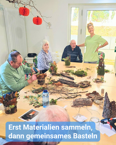 Seniorinnen und Senioren sitzen gemeinsam an einem großen Tisch mit einer Betreuungskraft. Sie nutzen Naturmaterialien, um Herbstdekoration zu gestalten. Bildbeschriftung: „Erst Materialien sammeln, dann gemeinsames Basteln.“