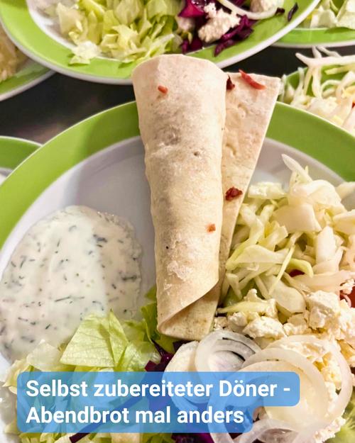 Nahaufnahme eines Tellers mit zwei Fladenbroten, möglicherweise Pita oder Tortillas, die so gefaltet sind, dass sie einem Döner Kebap ähneln. Diese sind eingebettet in eine lebendige Mischung aus zerrissenem Salat, fein gehackten Zwiebeln und offenbar zerbröckeltem Feta-Käse. An der Seite ist eine cremige, krautige Sauce sichtbar, vermutlich ein joghurtbasierter Dip.  Der Hintergrund zeigt unscharfe Elemente, die auf weitere Teller mit Speisen und möglicherweise eine Restaurantatmosphäre hindeuten. Ein Teller präsentiert einen Salat mit rötlich-violetten Gemüsesorten.  Eine über dem Bild eingeblendete Beschriftung lautet: "Selbst zubereiteter Döner - Abendbrot mal anders".