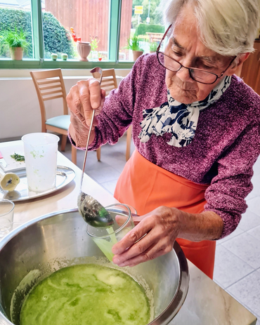 Eine ältere Frau steht vor einem großen Topf mit Gurkenlimonade. Sie schöpft die Speise vom Topf in ein Glas.
