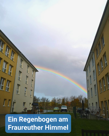 Zwischen zwei gelben Gebäuden ist der dunkle Himmel, geteilt von einem bunten Regenbogen, zu sehen. Bildbeschriftung: „Ein Regenbogen am Fraureuther Himmel“
