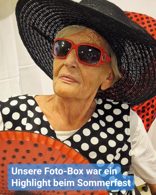 Eine ältere Frau posiert mit großem Hut, roter Sonnenbrille und Fächer vor schwarzem Punktmuster. Text: 'Unsere Foto-Box war ein Highlight beim Sommerfest'.
