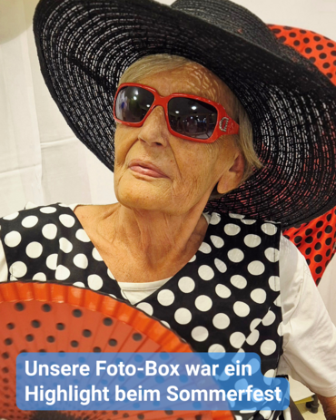 Eine ältere Frau posiert mit großem Hut, roter Sonnenbrille und Fächer vor schwarzem Punktmuster. Text: 'Unsere Foto-Box war ein Highlight beim Sommerfest'.