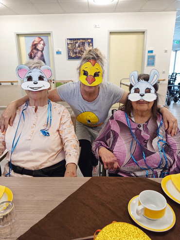 Zwei Seniorinnen mit Faschingsmasken sitzen an einem Tisch. Hinter ihnen ist eine Betreuungskraft,  die ebenfalls eine Maske trägt.