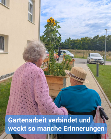 Eine ältere Frau und daneben ein älterer Herr im Rollstuhl betrachten gemeinsam das bunte Blumen-Hochbeet mit mit einer großen Sonnenblume in der Mitte. Bildbeschriftung: Gartenarbeit verbindet und weckt so manche Erinnerungen