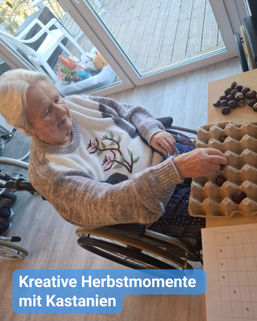 Eine Seniorin im Rollstuhl mit einem grauen Pullover sitzt an einem Tisch und legt Kastanien in einen Eierkarton nach einem Muster ab. Bildbeschriftung: „Kreative Herbstmomente mit Kastanien”
