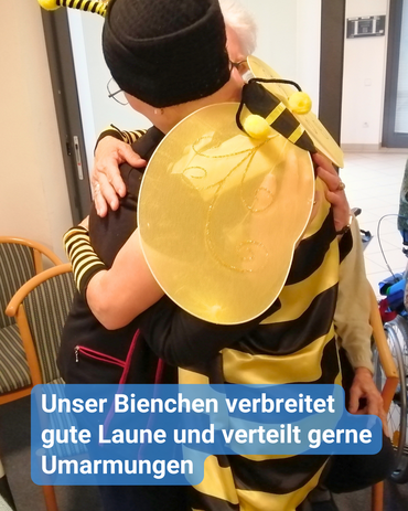 Eine Mitarbeiterin im Bienenkostüm umarmt eine Seniorin. Bildbeschriftung: „Unser Bienchen verbreitet gute Laune und verteilt gerne Umarmungen.“