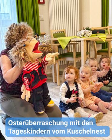 Eine Frau mit lockigem, braunem Haar sitzt auf einem Holzfußboden und hält eine Puppe in den Händen. Die Puppe ist ein fantasievoller Charakter mit langen, braunen Haaren, bekleidet mit einem rot-weiß gestreiften Shirt und einer dunklen Jeans. Die Frau trägt ein langärmliges, dunkelgraues Oberteil. Vor ihr sitzen mehrere junge Kinder, die aufmerksam zuschauen. Die Kinder sind unterschiedlichen Alters, haben verschiedene Haarfarben und tragen legere Kleidung. Sie scheinen vom Puppenspiel fasziniert zu sein. Der Schauplatz erinnert an einen Raum in einer Kindertagesstätte oder Vorschule. Im Hintergrund steht ein Tisch mit verschiedenen Gegenständen, was auf eine Lernumgebung hinweist. Eine hellfarbige Wand und grüne Vorhänge runden das Gesamtbild ab. Die Atmosphäre des Bildes ist warm und einladend. Der über das Bild gelegte Text lautet: "Osterüberraschung mit den Kindergartenkindern vom Kuschelnest" (Kuschelnest dürfte der Name der Einrichtung sein).