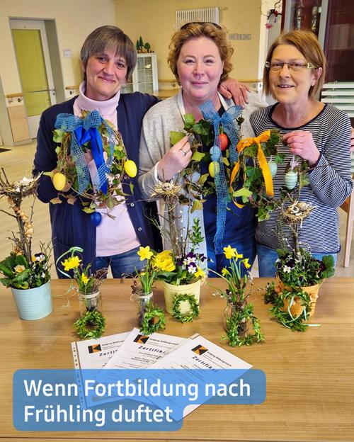 Drei Frauen stehen in einem Raum und lächeln in die Kamera. Sie halten frühlingshafte Bastelarbeiten, die kleine Arrangements aus Zweigen, Grünpflanzen und Ostereiern beinhalten. Diese Arrangements sind auf einem Tisch vor ihnen ausgestellt, zusammen mit weiteren kleinen dekorativen Gegenständen in Gläsern und Töpfen. Auf dem Tisch liegen zudem Zertifikate, die darauf hindeuten, dass die Frauen eine Art Kurs oder Schulung abgeschlossen haben.  Die Frauen sind lässig gekleidet. Die Dame ganz links trägt einen mauvefarbenen Pullover. Die Frau in der Mitte präsentiert ein türkisfarbenes Shirt. Die Frau ganz rechts trägt ein gestreiftes Langarmshirt. Der Hintergrund ist leicht verschwommen, zeigt jedoch einen hellen Raum mit sichtbaren Möbeln und Dekoration.  Am unteren Rand des Bildes befindet sich ein Spruch: "Wenn Fortbildung nach Frühling duftet." Der Text ist in einem blauen Banner abgedruckt.