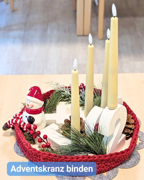 Ein selbst gestalteter Adventskranz steht auf einem Tisch: Vier hohe, cremefarbene Kerzen, Tannenzweige, Zapfen und rote Beeren sind in einem gestrickten roten Rahmen arrangiert. Daneben sitzt eine kleine Schneemannfigur. Die Dekoration wirkt festlich und zeigt ein kreatives Adventskranzbinden in einer Pflegeeinrichtung.