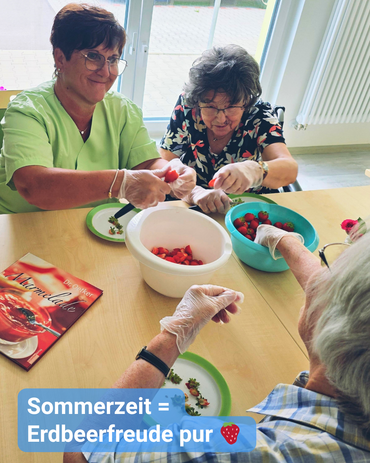 Drei Personen, darunter eine Pflegekraft, bereiten Erdbeeren am Tisch zu. Text: 'Sommerzeit = Erdbeerfreude pur'.