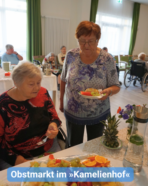 Zwei ältere Frauen, eine davon im Rollstuhl, wählen verschiedenes Obst an einem Buffettisch aus. Im Hintergrund sind viele Senioren an Tischen zu sehen. Bildbeschriftung: Obstmarkt im »Kamelienhof«