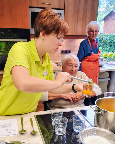 Eine Seniorin im Rollstuhl sitzt vor der Herdplatte und schaut in den Kochtopf, während einer Pflegekraft die Speise in ein Glas gibt.