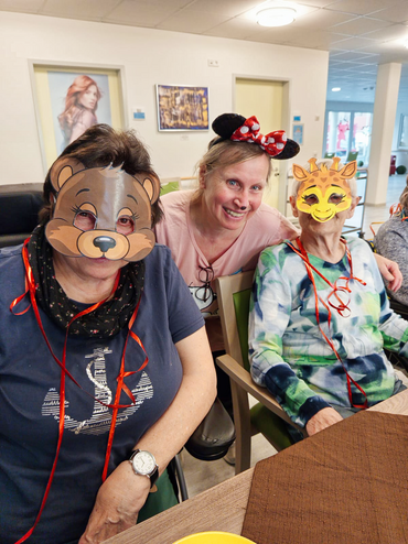 Zwei Seniorinnen mit Faschingsmasken sitzen an einem Tisch. Hinter ihnen ist eine Betreuungskraft, mit Minnie Maus Ohren.