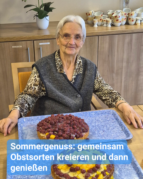 Ältere Dame sitzt an einem Tisch und lächelt freundlich in die Kamera. Vor ihr stehen zwei runde Obstkuchen, ganz frisch, gebacken und dekoriert. Bildunterschrift: “Sommergenuss: gemeinsam Obstsorten kreieren und dann genießen