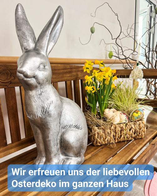 Eine silberne Kaninchenstatue thront auf einer hölzernen Bank. Das große Kaninchen schaut nach links. Sein Fell ist strukturiert und wirkt metallic.  Hinter dem Kaninchen befindet sich ein geflochtener Korb, gefüllt mit gelben Narzissen, grünem Gras sowie kleinen dekorativen Ostereiern und -kaninchen. Oberhalb des Korbes hängt ein kahler, dunkelbrauner Zweig, an dem kleine dekorative Eier baumeln.  Die Bank besteht aus dunkelbraunem Holz und scheint Teil eines größeren Arrangements zu sein.  Am unteren Bildrand prangt ein hellblaues Banner mit dem Text: "Wir erfreuen uns der liebevollen Osterdeko im ganzen Haus."