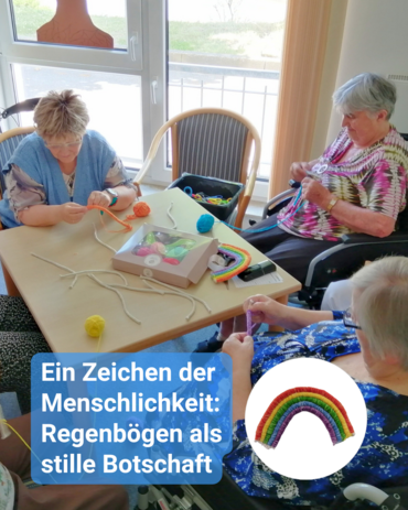 Drei Frauen häkeln am Tisch bunte Regenbögen. Zu sehen eine Kiste mit Bastelmaterial. Unten rechts ein gehäkelter Regenbogen. Text: 'Ein Zeichen der Menschlichkeit: Regenbögen als stille Botschaft'.