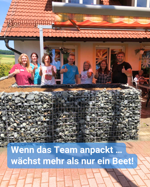 Mehrere Mitarbeiter stehen lächelnd hinter einem großen Hochbeet aus Gabionen. Text: 'Wenn das Team anpackt ... wächst mehr als nur ein Beet!'.