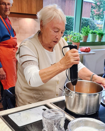 Im Vordergrund ist eine Seniorin am Kochtopf, sie püriert den Inhalt.