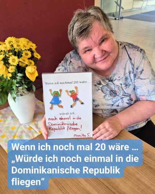 Eine ältere Frau sitzt an einem Holztisch, welcher mit einer Vase und gelben Blümchen dekoriert ist. Sie hält einen Zettel in die Kamera. Bildbeschriftung: Wenn ich noch mal 20 wäre … „Würde ich noch einmal in die Dominikanische Republik fliegen“