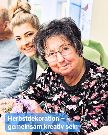 An einem großen Tisch mit Herbstdekoration sitzen ältere Menschen, und zwischen ihnen lächelt eine Betreuungskraft in die Kamera. Bildbeschriftung: Herbstdekoration – gemeinsam kreativ sein