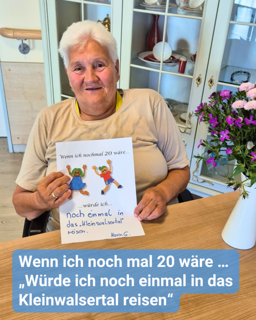 Eine ältere Dame sitzt an einem Holztisch, welcher mit einem bunten Blumenstrauß dekoriert ist, und hält einen Zettel in die Kamera. Bildbeschriftung: Wenn ich noch mal 20 wäre … „Würde ich noch einmal in das Kleinwalsertal reisen“