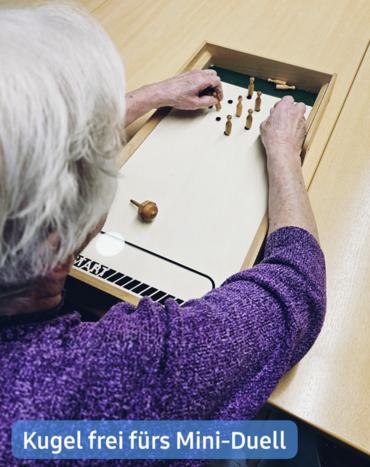 Nahaufnahme eines älteren Menschen mit kurzen, weißen Haaren, der einen lila Pullover trägt und ein Tischspiel spielt.  Das Spiel scheint eine Art Miniatur-Kegeln oder etwas Ähnliches zu sein, mit kleinen Holzklötzen und einer Kugel (oder möglicherweise einer Murmel), die auf einer weißen Spielfläche sichtbar sind. Das Spielbrett ist in einen Holztisch eingelassen.  Die Person ist aktiv in das Spiel vertieft und bewegt geschickt die Spielstücke mit ihren Händen. Der Fokus liegt scharf auf dem Spiel und den Händen der Person, während der Hintergrund sanft verschwommen ist. Eine Bildunterschrift am unteren Rand lautet: "Kugel frei fürs Mini-Duell."