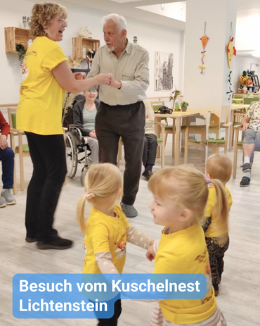 Tanz mit Jung und Alt. Während im Hintergrund ein älteres Pärchen tanzt, sieht man im Vordergrund drei kleine Kinder, die sich an den Händen fassen und im Kreis tanzen. Bildbeschriftung: Besuch vom Kuschelnest Lichtenstein