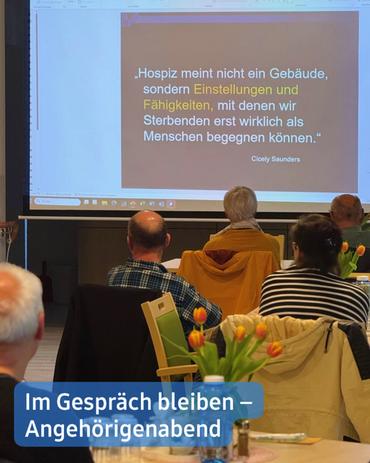 Eine Projektionsleinwand zeigt ein Zitat von Cicely Saunders über die Palliativversorgung, das betont, dass es sich dabei nicht lediglich um ein Gebäude handelt, sondern um eine Reihe von Haltungen und Fertigkeiten, die eine bedeutungsvolle Verbindung zu den Sterbenden ermöglichen.  Im unteren Bereich des Bildes ist eine Gruppe von Menschen zu sehen, die in Stühlen sitzen und während eines Meetings oder einer Präsentation der Leinwand zugewandt sind. Die meisten blicken auf den Bildschirm, während einige in Gespräche vertieft sind. Ein Blumenstrauß steht im Vordergrund auf dem Tisch. Eine Titelkarte am unteren Rand weist den Text "Im Gespräch bleiben - Angehörigenabend" auf, was übersetzt "Im Gespräch bleiben - Angehörigenabend" bedeutet.  Die insgesamt düstere, jedoch hoffnungsvolle Atmosphäre deutet auf ein Treffen hin, das möglicherweise einer Unterstützungsgruppe oder einer Bildungsveranstaltung zu den Themen Palliativversorgung und Trauer gewidmet ist. Die Beleuchtung ist gedämpft.
