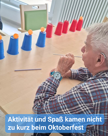 Auf einem Tisch sind umgedrehte Trinkbecher hingestellt, auf denen kleine Bälle platziert sind. Ein Senior soll mit einem Trinkhalm die Bälle wegpusten. Bildbeschriftung: Aktivität und Spaß kamen nicht zu kurz beim Oktoberfest.