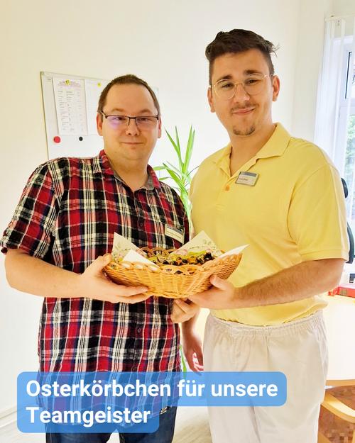 Zwei Männer stehen nebeneinander, lächeln und halten einen Korb aus Weide, der mit Süßigkeiten gefüllt ist.  Der Mann links trägt ein rot-schwarz kariertes Hemd, während der Mann rechts ein gelbes Poloshirt und weiße Hosen anhat. Beide blicken direkt in die Kamera.  Am unteren Rand des Bildes ist eine Bildunterschrift eingeblendet: "Osterkörbchen für unsere Teamgeister". Der Hintergrund zeigt einen hellen Raum mit einem sichtbaren Teil eines Whiteboards sowie einer Topfpflanze zwischen den beiden Männern. Zudem sind ein Teil eines Fensters und eine weiße Wand zu sehen.