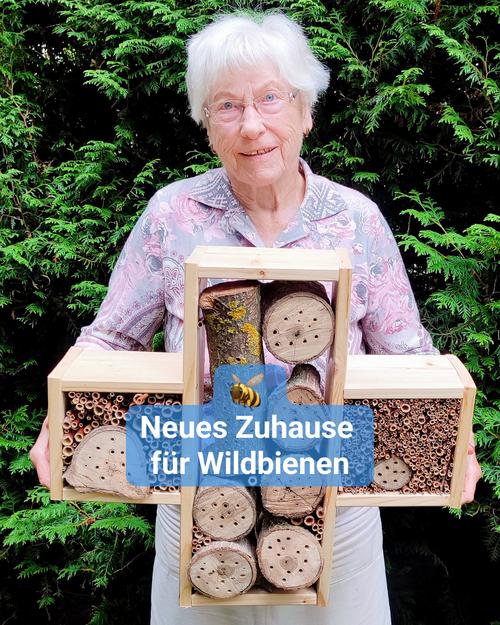 Eine Seniorin steht draußen vor einer grünen Hecke und hält ein großes Insektenhotel in den Händen. Bildbeschriftung: „Neues Zuhause für Wildbienen“