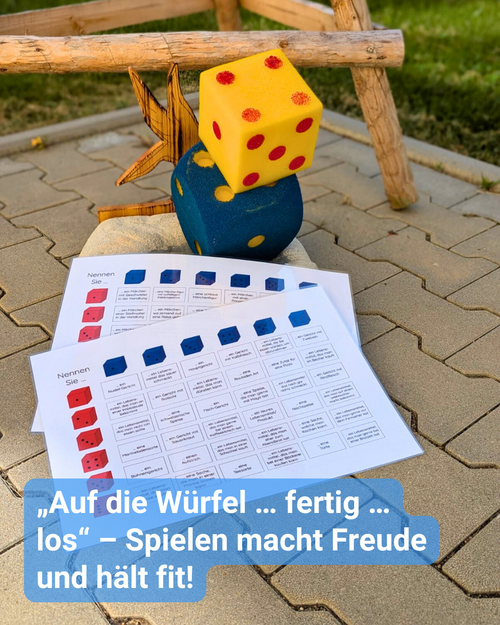 Draußen im Innenhof auf dem Pflaster liegen zwei große XXL Würfel in dunkelblau und gelb, nebst Spielanleitung. Bildbeschriftung: „Auf die Würfel … fertig … los“ – Spielen macht Freude und hält fit!