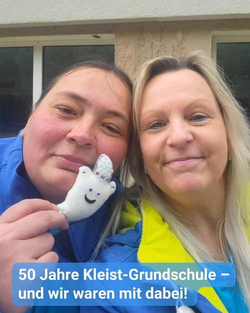 Ein Selfie von zwei Frauen im Freien vor eine beige Ziegelmauer mit zwei Fenstern mit weißen Rahmen im Hintergrund. Die Frau auf der linken Seite trägt eine blaue Jacke und hält ein kleines weißes Plüschspielzeug hoch, das einer Handpuppe ähnelt. Es ist ein lächerlicher Geist, welcher an der Spitze mit Glitzersteinchen verziert ist. Die Frau auf der rechten Seite trägt eine gelbe und blaue Jacke.  Beide Frauen lächeln und schauen in die Kamera. Am unteren Bildrand ist der Text überlagert: „50 Jahre Kleist-Grundschule – und wir waren mit dabei!“