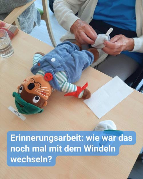 Eine Person sitzt an einem Tisch und wechselt die Windel eines Stoffotters.    Der Otter liegt auf dem Rücken auf dem holzfarbenen Tisch, trägt eine blaue Latzhose und eine grüne Kappe.    Die wechselnde Person trägt eine weiße Strickjacke über einem blauen Hemd.    Auf dem Tisch sind ein Wasserglas, eine Serviette und ein Zettel sichtbar.    Der Text auf dem Bild lautet: „Erinnerungsarbeit: wie war das noch mal mit dem Windeln wechseln?“