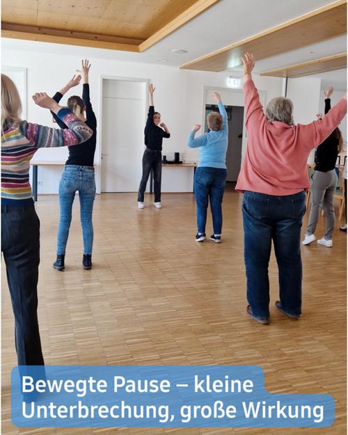Auf Augenhöhe betrachtet man eine Gruppe von Menschen, die an einer kurzen Übungseinheit oder einem Stretching teilnehmen, in einem Raum, der vermutlich ein Gemeindezentrum oder einen ähnlichen Ort darstellt.  Der Raum verfügt über lichtfarbene Holzböden und Wände sowie eine schlichte, unauffällige Decke. Die Teilnehmerinnen, überwiegend Frauen unterschiedlichen Alters, sind in lässiger Kleidung gekleidet. Sie führen sanfte Dehnübungen und Bewegungen aus, während ihre Arme erhoben sind und ihre Körper sich leicht neigen.  Am unteren Rand des Bildes ist ein Banner: „Aktive Pause – kleine Unterbrechung, große Wirkung.“